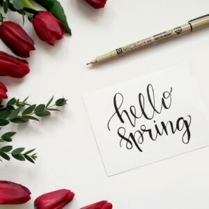 Close-up of red tulips beside a hand-lettered 'hello spring' card on white background.